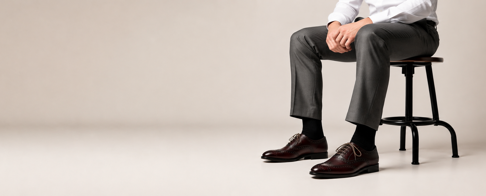Man seated on stool wearing grey trousers and burgundy leather brogue oxfords with contrast laces