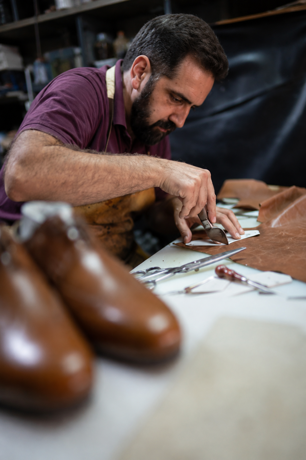 Craftsman hand-cutting leather with a knife at a workbench, with finished brown shoes in foreground