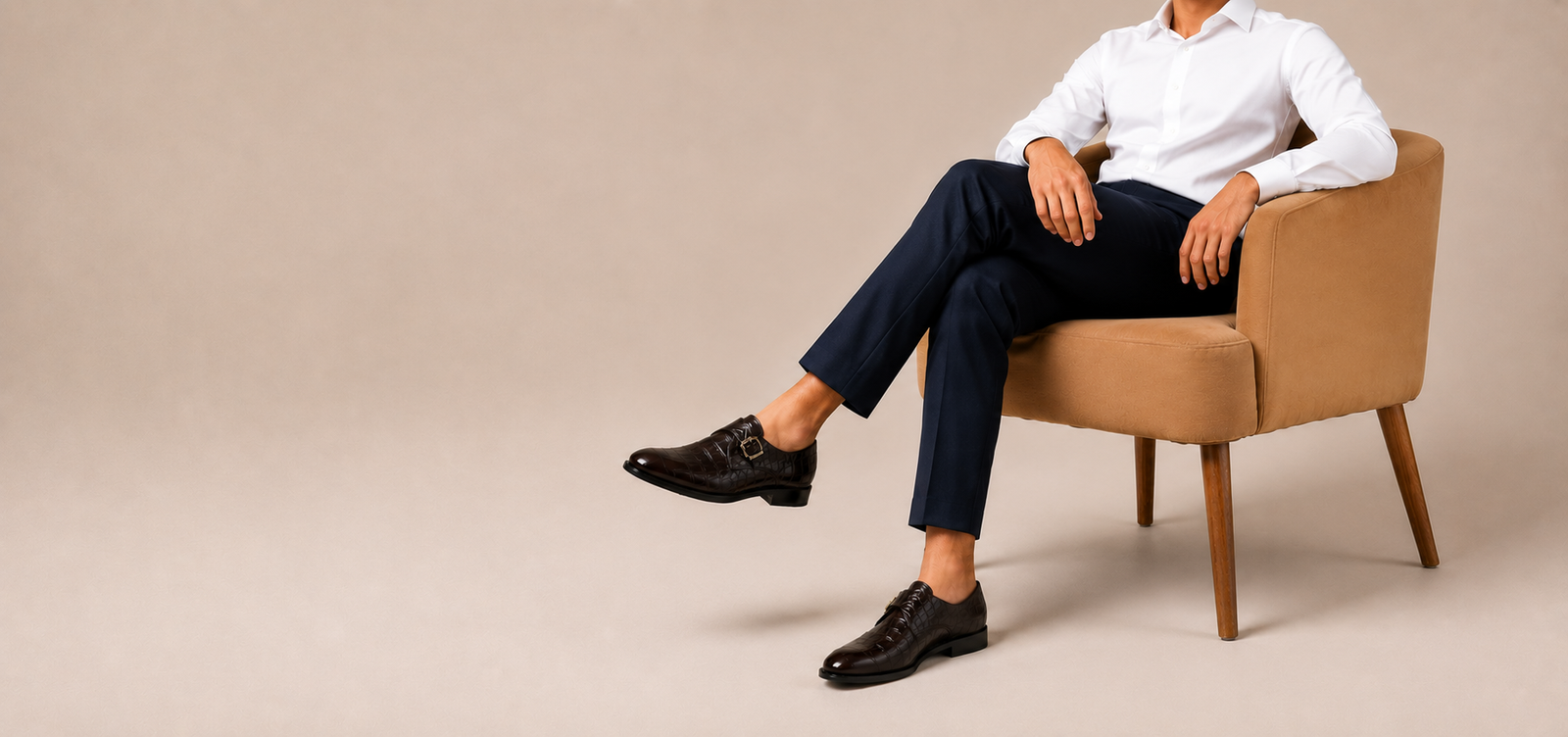Man seated in tan armchair wearing navy trousers and dark brown crocodile-embossed monk strap shoes
